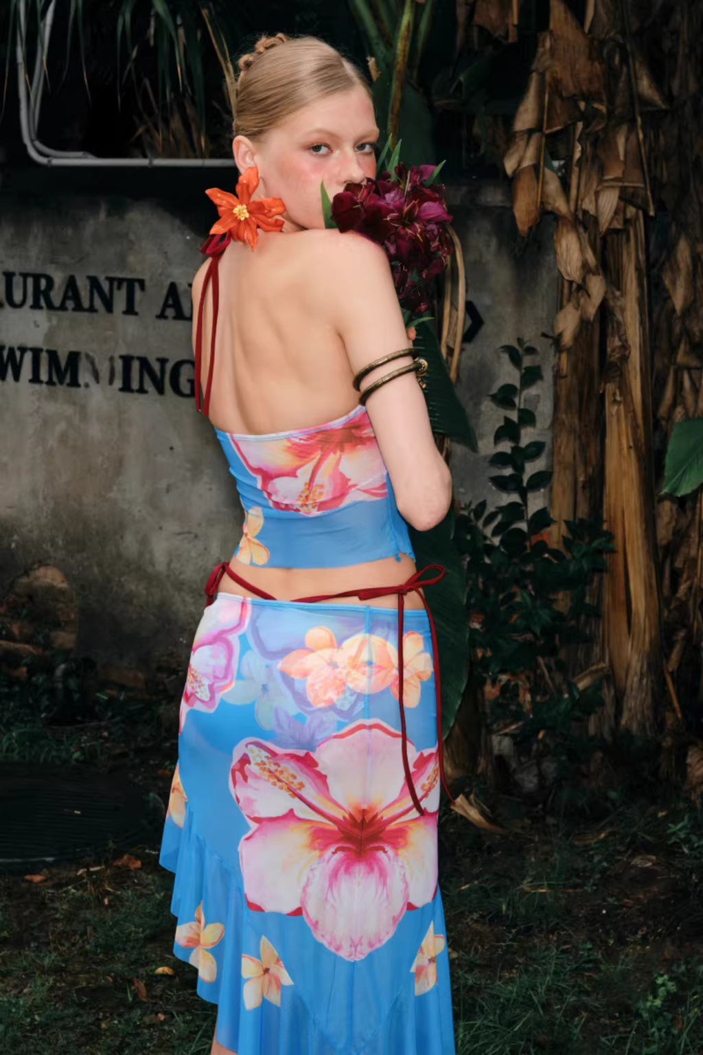 Woman in a floral dress standing outdoors with plants and a wall in the background