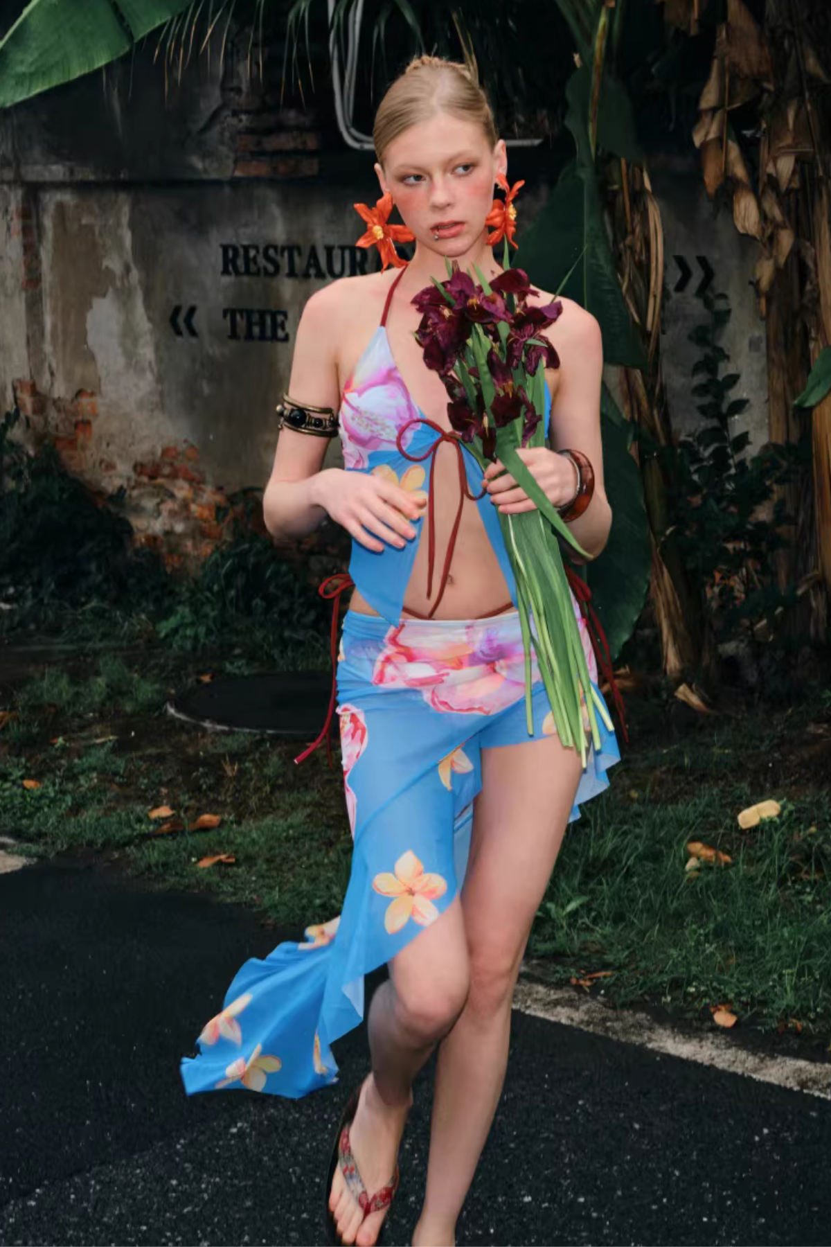 Woman in a colorful dress holding flowers against a rustic wall and plant background