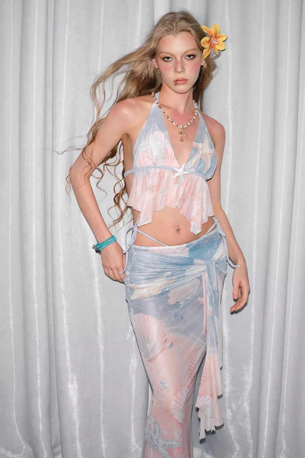 Model wearing a sheer, pastel-colored outfit with a floral headpiece against a white curtain background.