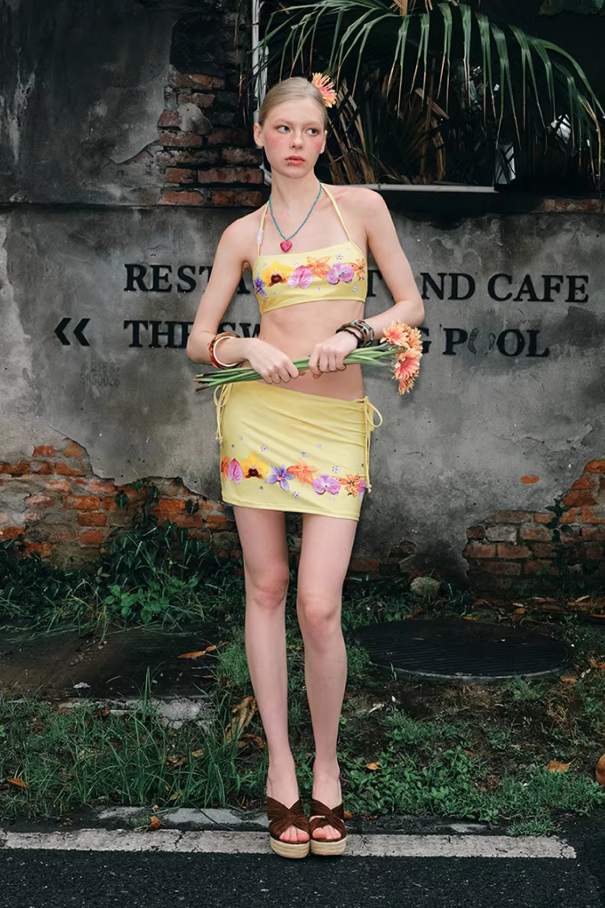 Woman in floral top and skirt standing in front of a rustic wall with text.