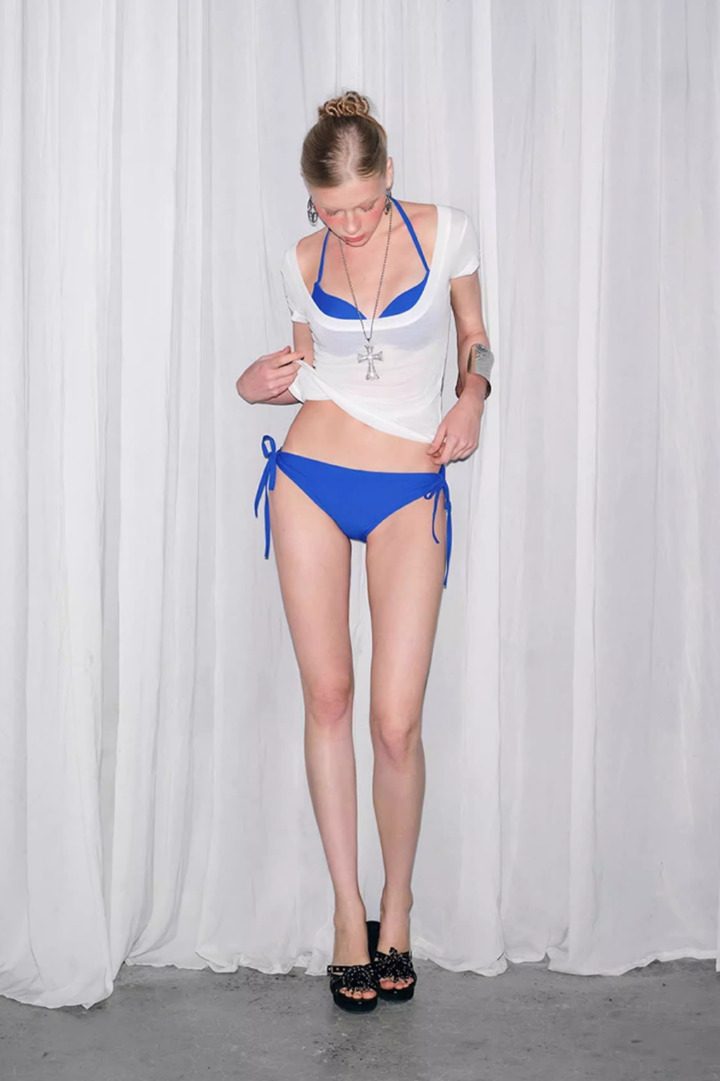 Woman wearing a white top and blue bikini bottom against a white curtain background